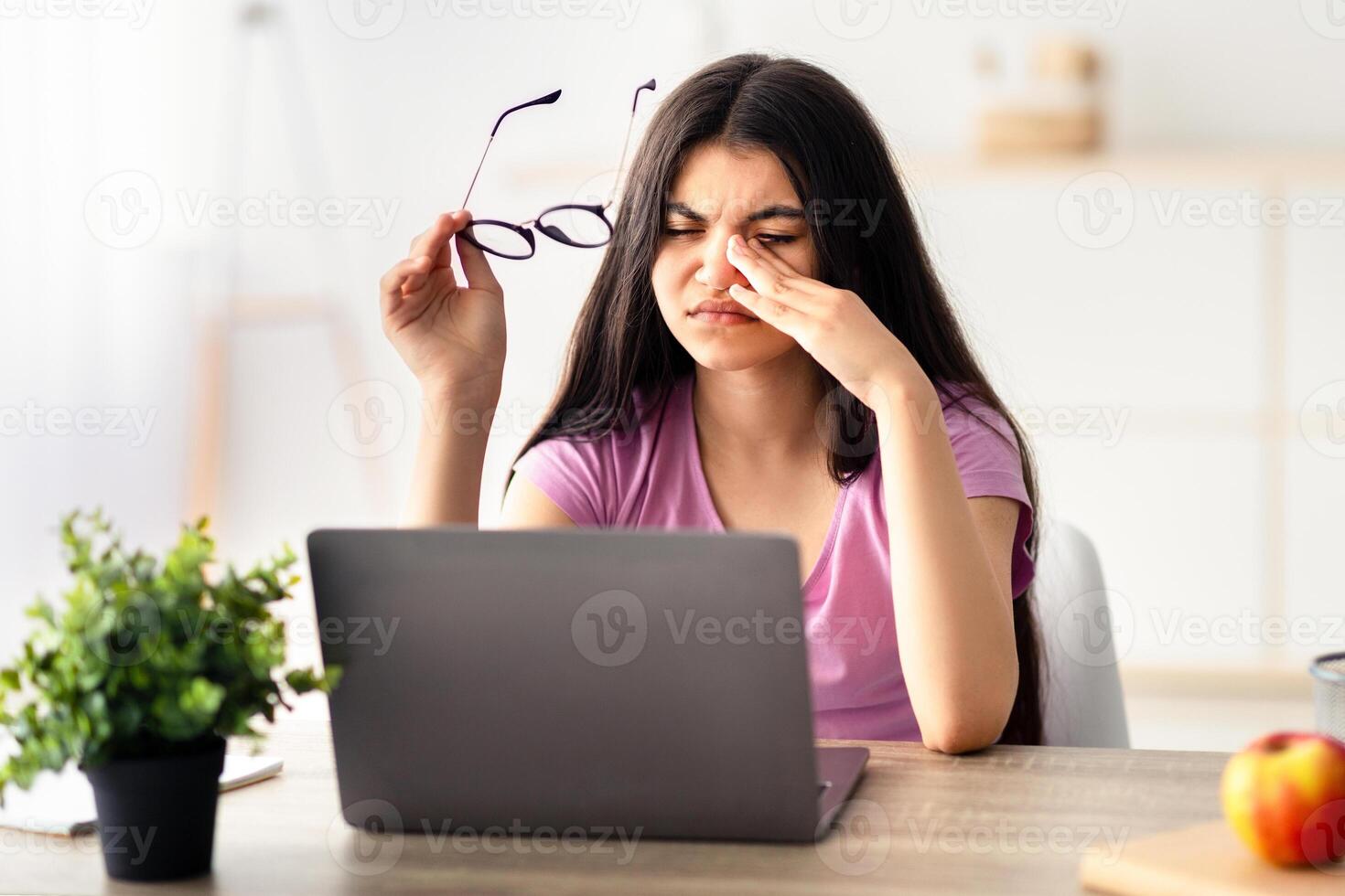 Indian teenage girl feeling tired, rubbing irritated eyes, sitting at desk with laptop, exhausted from online studies. Overworked adolescent having visual fatigue, doing home assignment on web photo