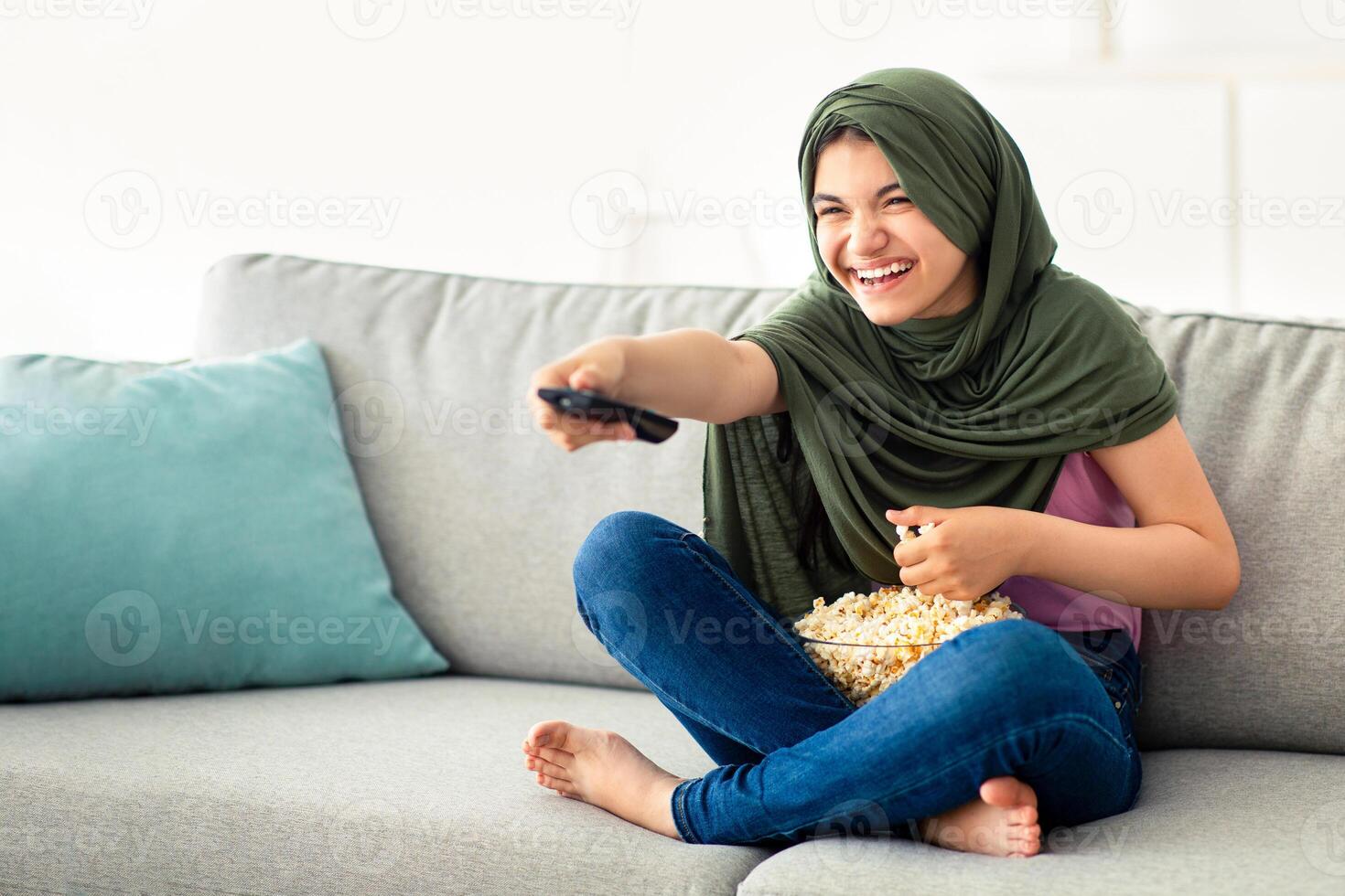 Domestic entertainments. Happy Muslim teenage girl in hijab with TV controller enjoying movie and eating popcorn indoors. Happy adolescent in traditional headscarf watching television on couch at home photo