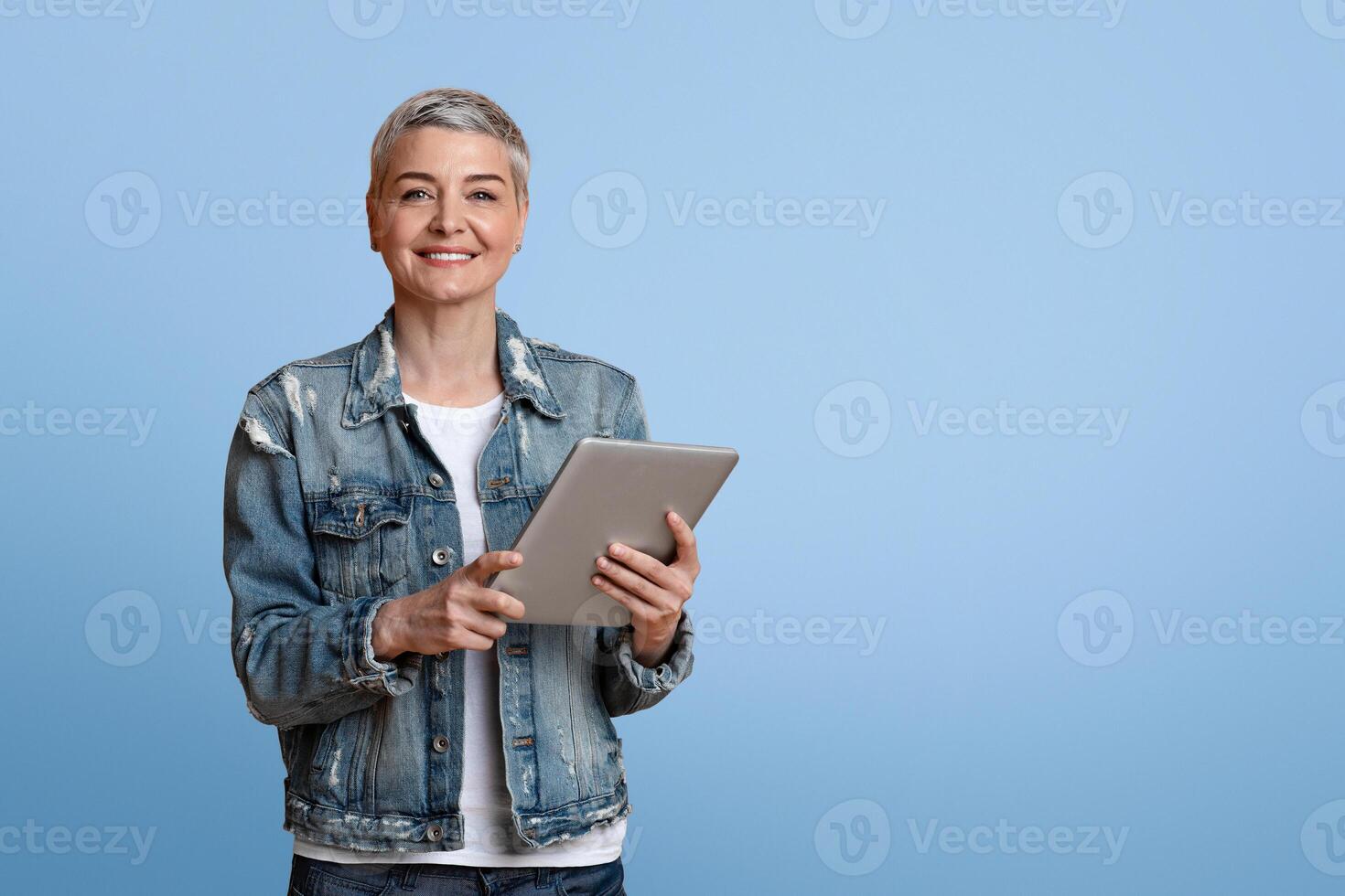 Modern Seniors. Portrait of smiling mature woman with digital tablet in hands posing on yellow background and looking to camera, copy space photo