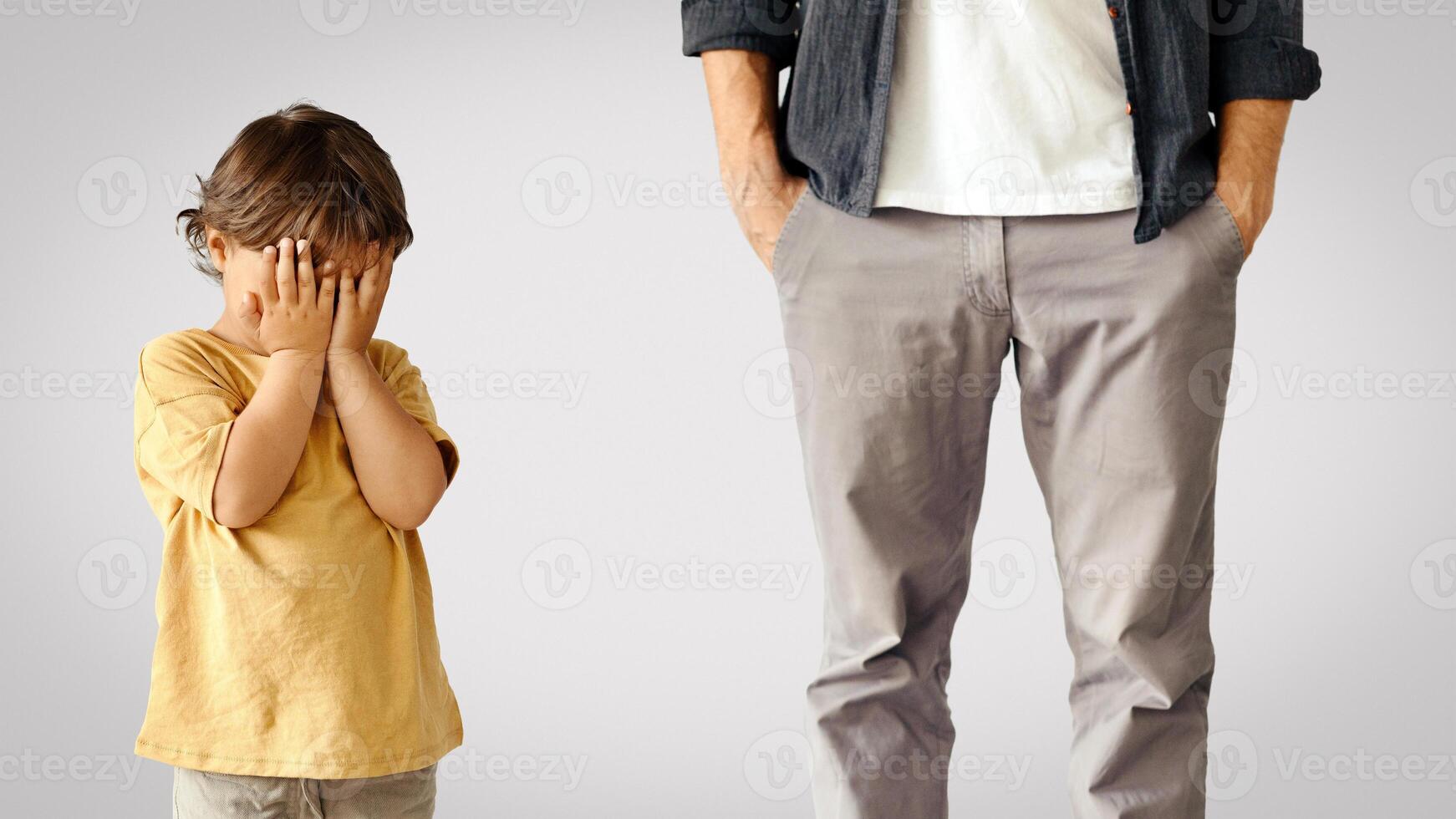 Kids offense. Little baby boy crying, feeling abandoned and lonely, unrecognizable father standing behind with hand in pockets, ignoring unhappy child, white background photo