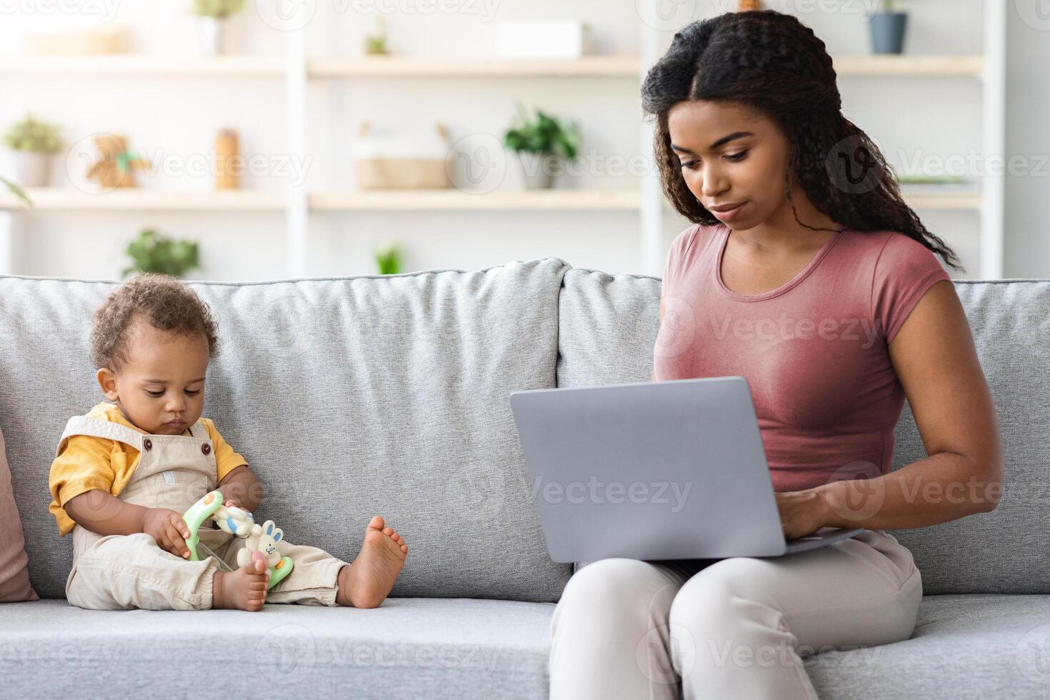 Young Mom Working Online On Computer With Little Baby Playing Around, Portrait Of African American Mother Freelancing On Laptop And Taking Care About Her Infant Son At Home, Free Space photo
