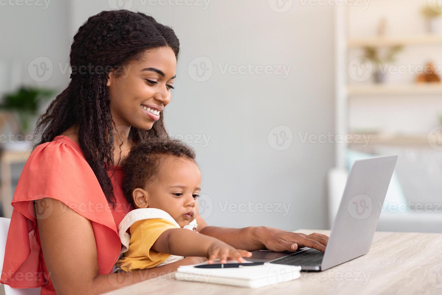 Portrait Of Young Woman With Baby On Hands Using Laptop At Home, Happy Smiling African American Lady Study Or Working Remotely On Computer While Looking After Her Child, Side View, Free Space photo