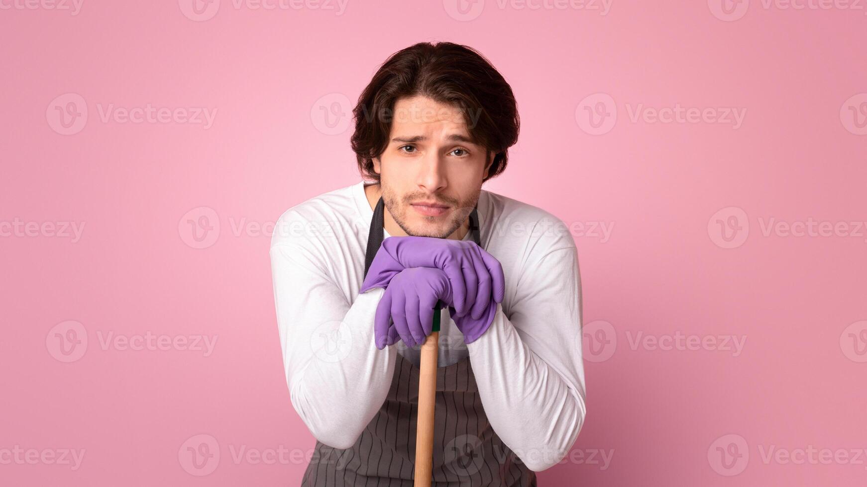 Tired Man Leaning At Mop Handle, Resting Head On Hands Exhausted After Spring Cleaning, Standing Over Pink Background With Free Space photo