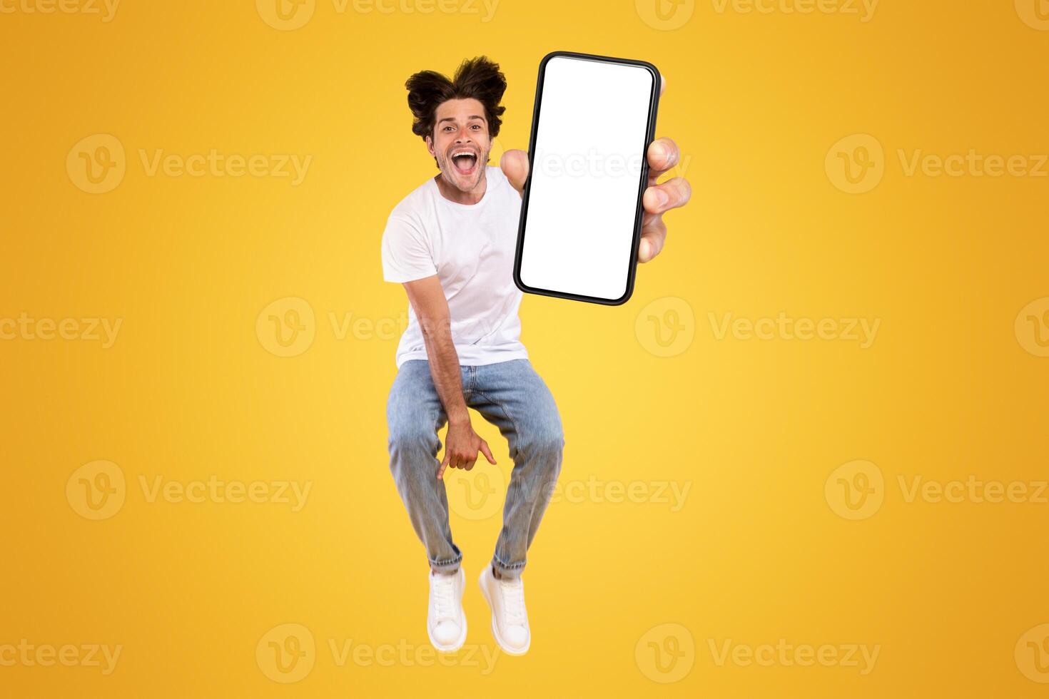 Handsome bearded young man recommending new mobile application, vertical collage. Excited guy holding smartphone, showing white blank screen, jumping up over orange yellow studio background photo