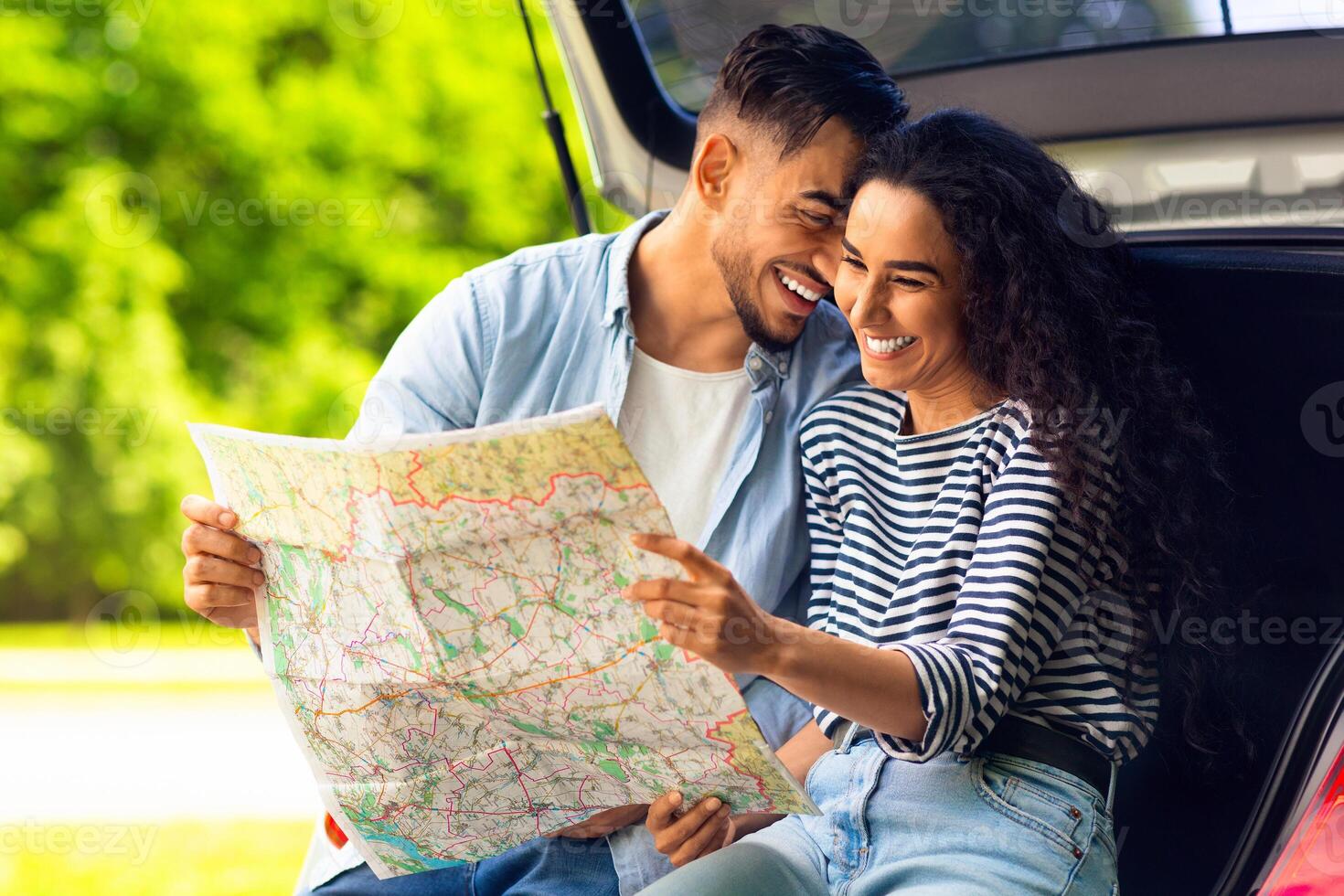 Happy young lovers cuddling while sitting on auto trunk and watching road map, copy space. Handsome arab guy bonding with his beautiful girlfriend, having break while car trip together photo