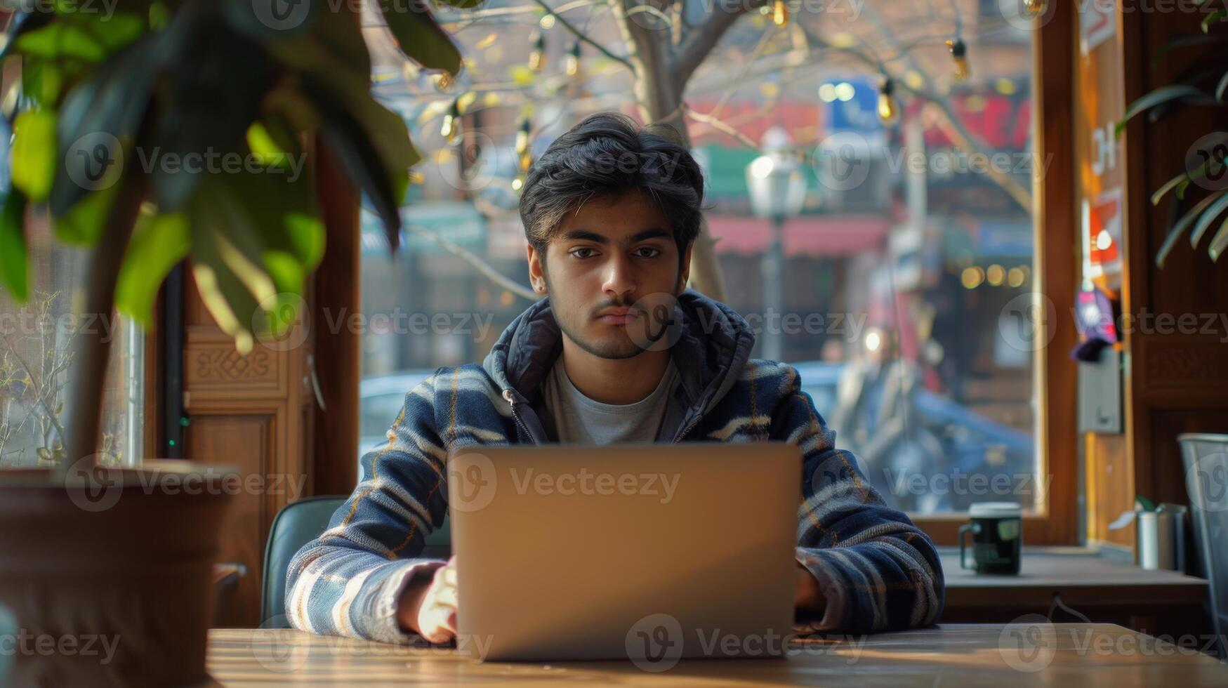 A young man with a serious expression works intently on his laptop at a cafe table. Sunlight pours in, illuminating the warm atmosphere of the space. photo