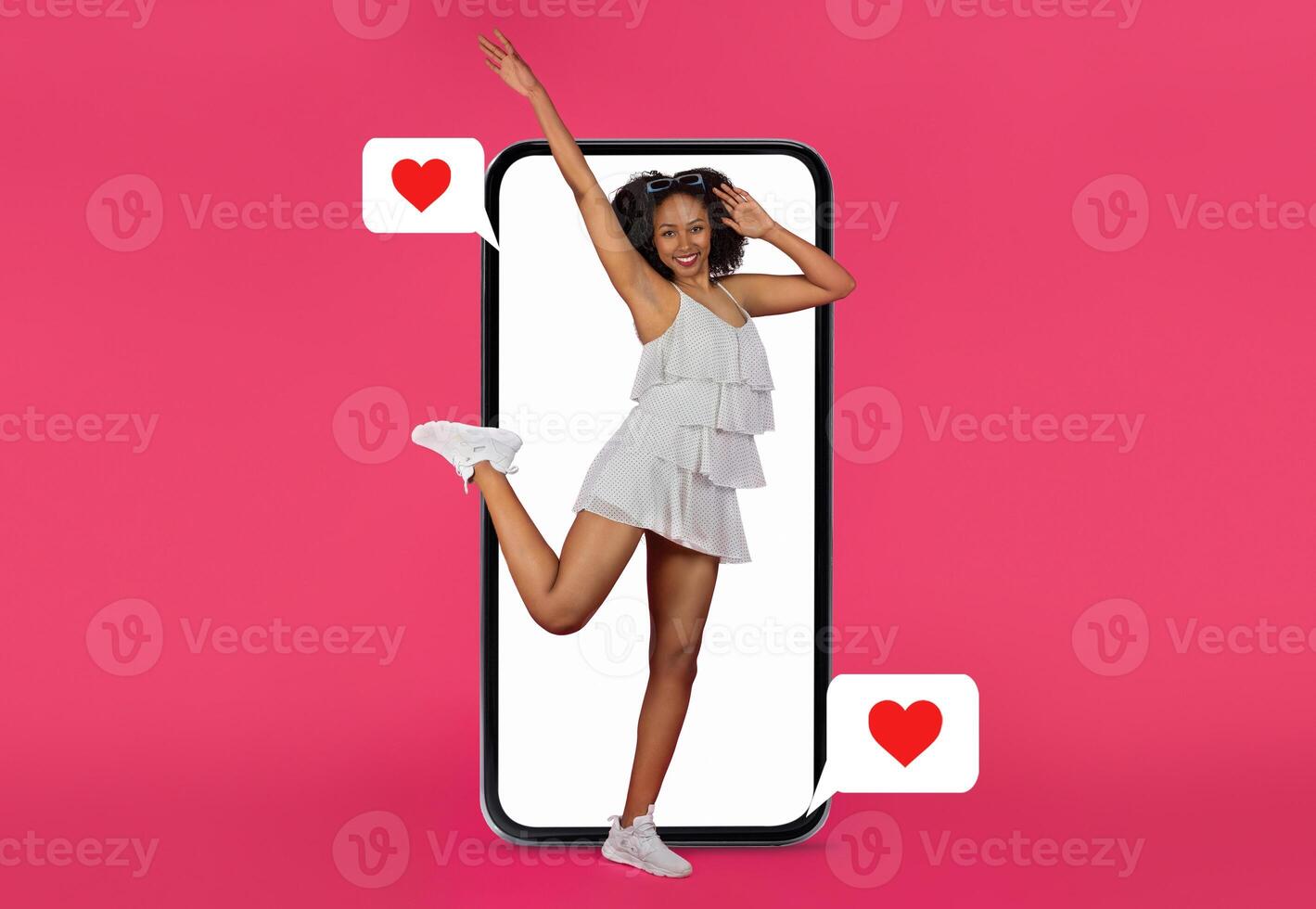 A young woman in a white dress strikes a playful pose with a smartphone. She expresses joy and excitement, holding a heart symbol, showcasing the connection of dating and social media. photo