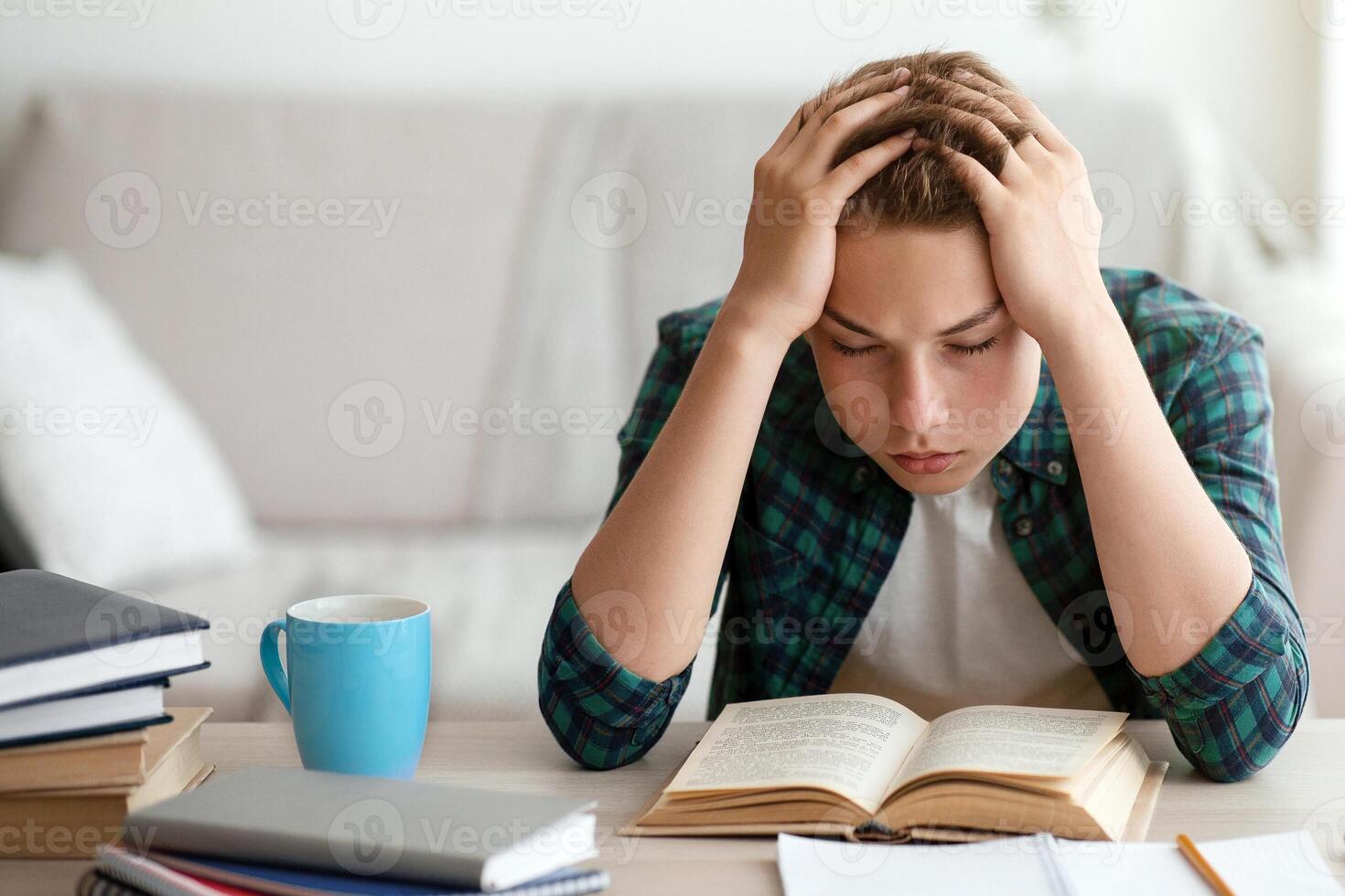 Exhausted teenager studying at home, getting ready before exams, reading book and using laptop, copy space photo