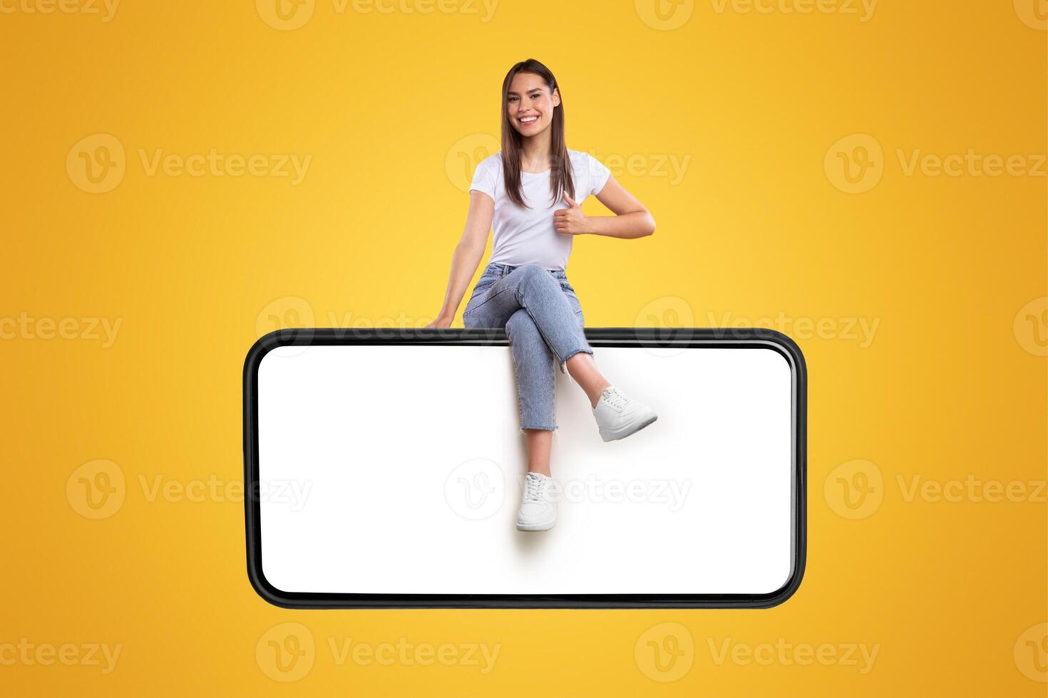 Cheerful young woman sitting on big huge horizontal new cellphone with white empty screen and showing thumb up gesture, mockup, casual lady enjoying newest entertaining mobile app, orange wall photo