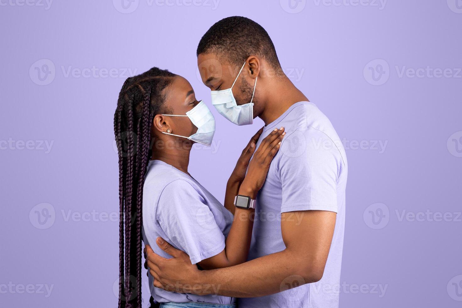 Romance During Pandemic Time. Side view profile portrait of smiling African American couple in medical face masks hugging, enjoying time together, standing isolated over purple studio background photo