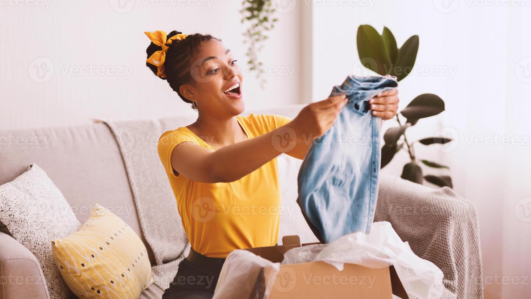 Delivery Concept. Smiling black woman holding new jeans, unboxing cardboard package, sitting on the couch in living room at home, free space. Excited lady happy with purchase and clothes shipment photo