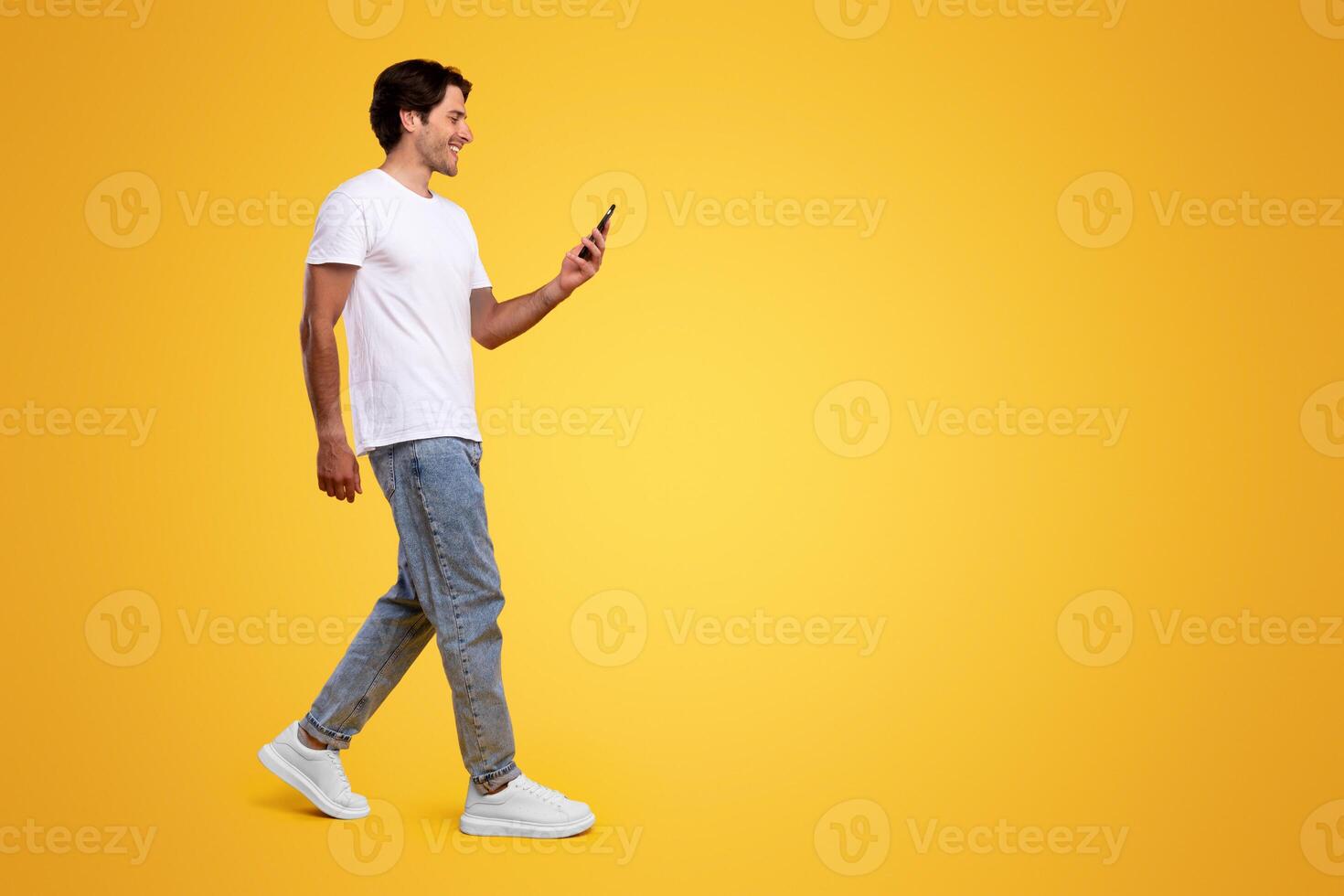 Communication, Technology And Lifestyle Concept. Full body length of smiling young guy using cell phone, walking isolated on orange studio background, profile side view panoramic banner, free space photo