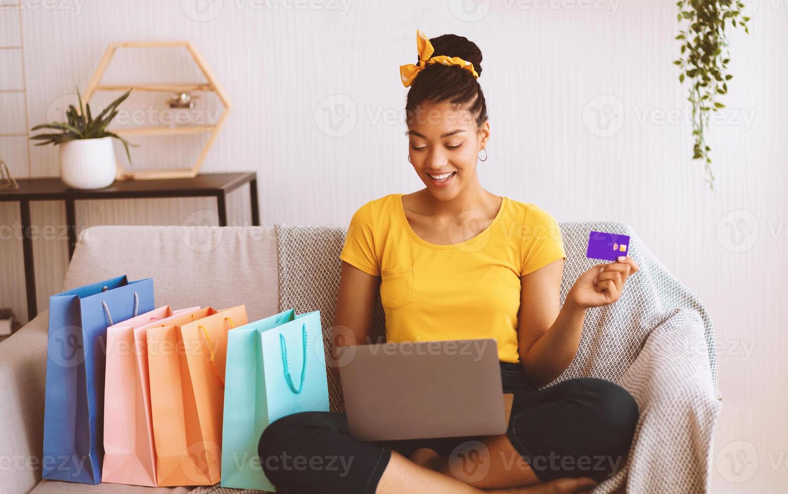 Online Payment Concept. Smiling african american lady holding credit card and using laptop, ordering goods and purchases via internet, sitting on the sofa with colorful shopping bags in living room photo