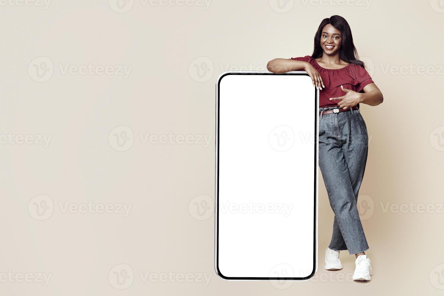Stylish millennial black woman pointing at big mobile phone with white blank display, mockup, happy african american lady in casual recommending newest mobile application, beige studio background photo
