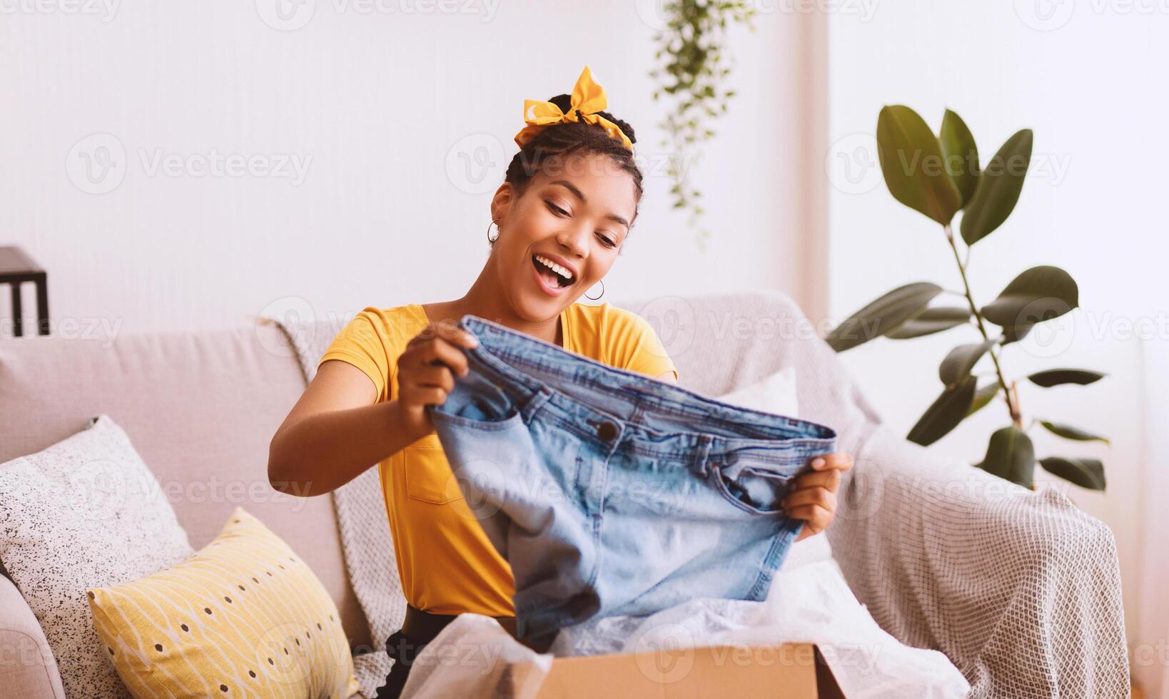 Delivery Concept. Happy black woman sitting on couch and holding jeans, unboxing cardboard package, free space photo