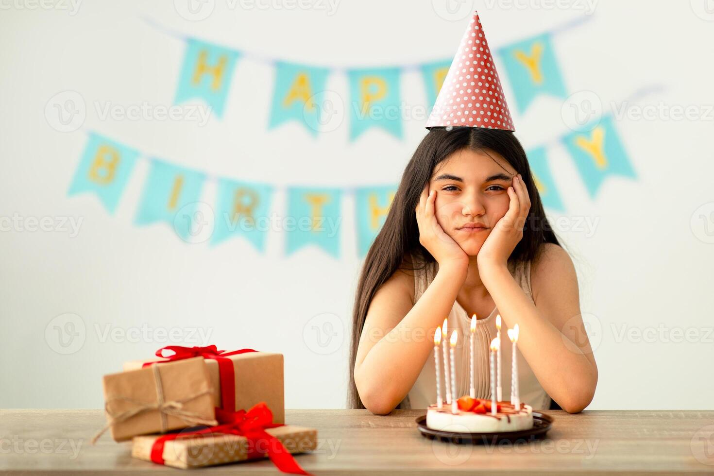 Portrait of disappointed Indian teen girl in party hat sitting alone at table with gifts and birthday cake, looking at camera, celebrating special day by herself, feeling unhappy and stressed photo
