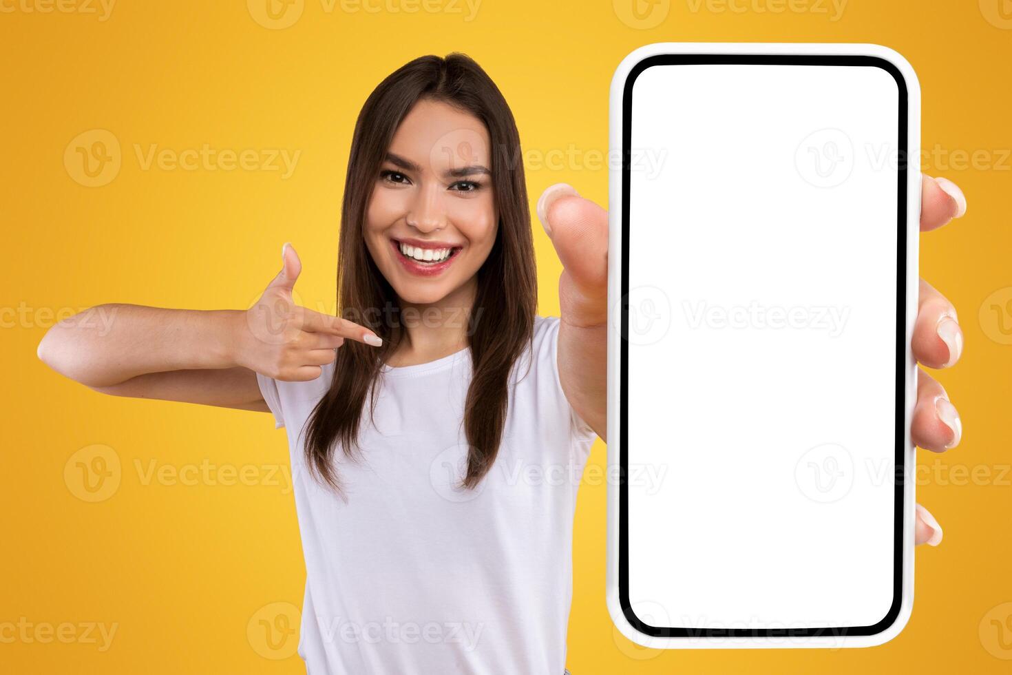 Check This Website. Portrait of smiling female holding smartphone with white blank screen in hand, showing device close up to camera and pointing finger at gadget with empty free space for mock up photo