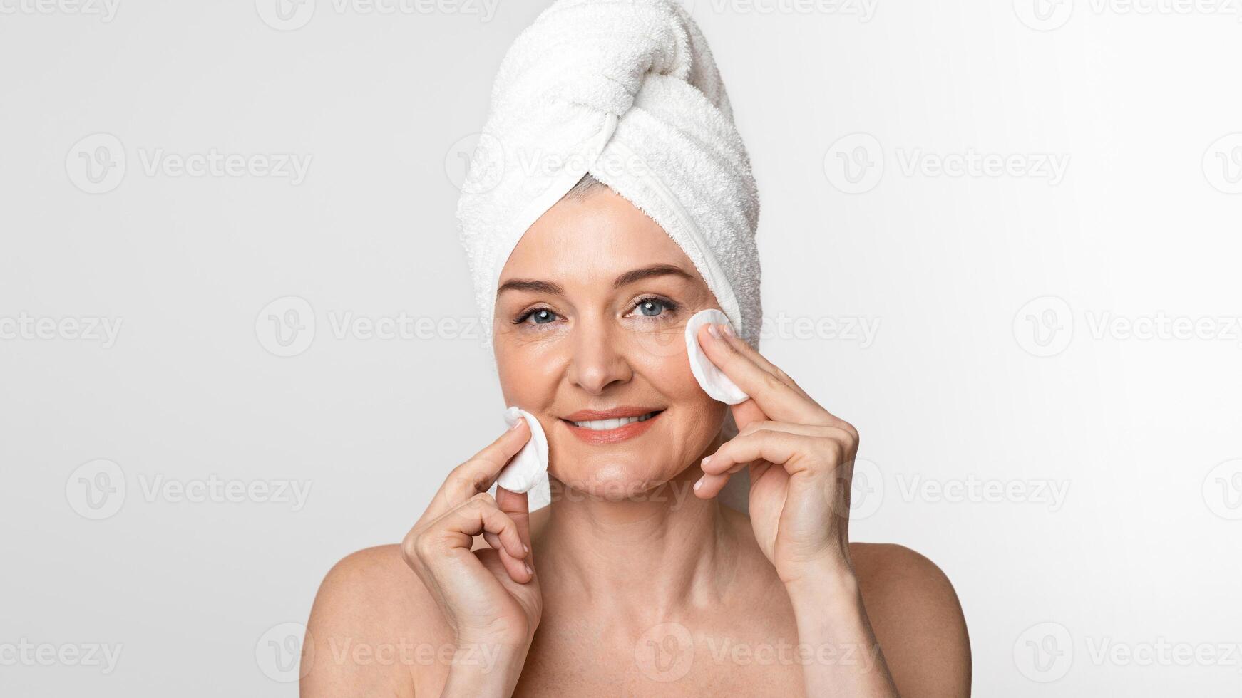 Skincare in mature age. Beautiful middle aged woman with towel on head cleaning her face with toner and cotton pads and looking at camera, copy space photo