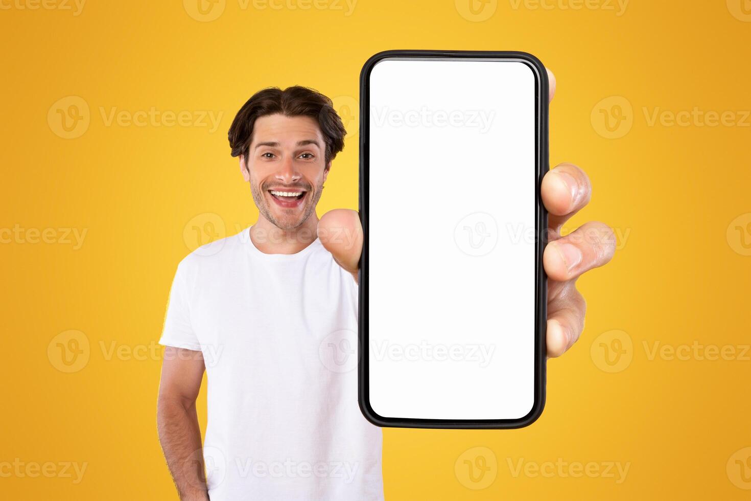 Mobile App Advertisement. Handsome Excited Man Showing White Empty Smartphone Screen Recommending App Posing Over Orange Studio Background, Smiling To Camera. Check This Out, Cellphone Display Mock Up photo