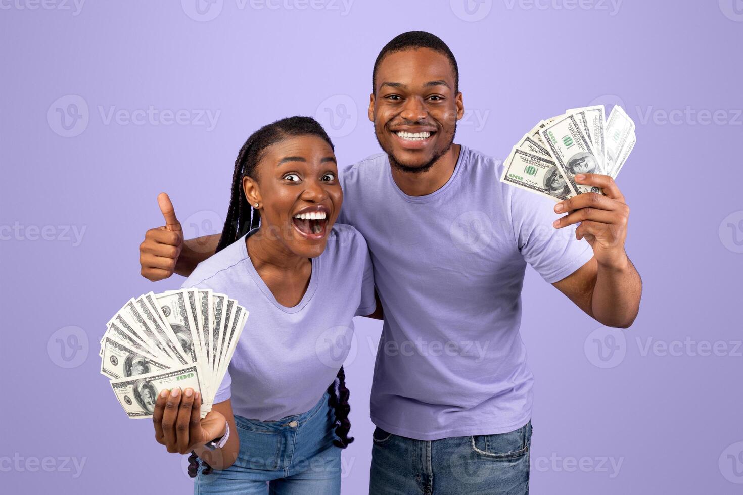 Portrait of overjoyed young African American couple holding and showing bunch of money, celebrating success together, making thumb up gesture, posing over purple studio background, copy space photo