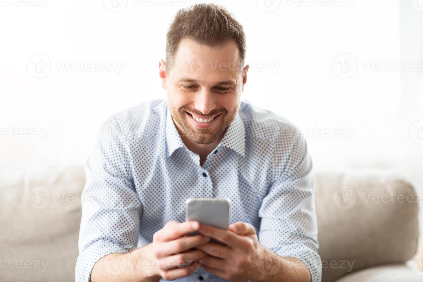 Messaging At Social Network. Happy millennial guy using modern smartphone device, sitting on sofa at home photo