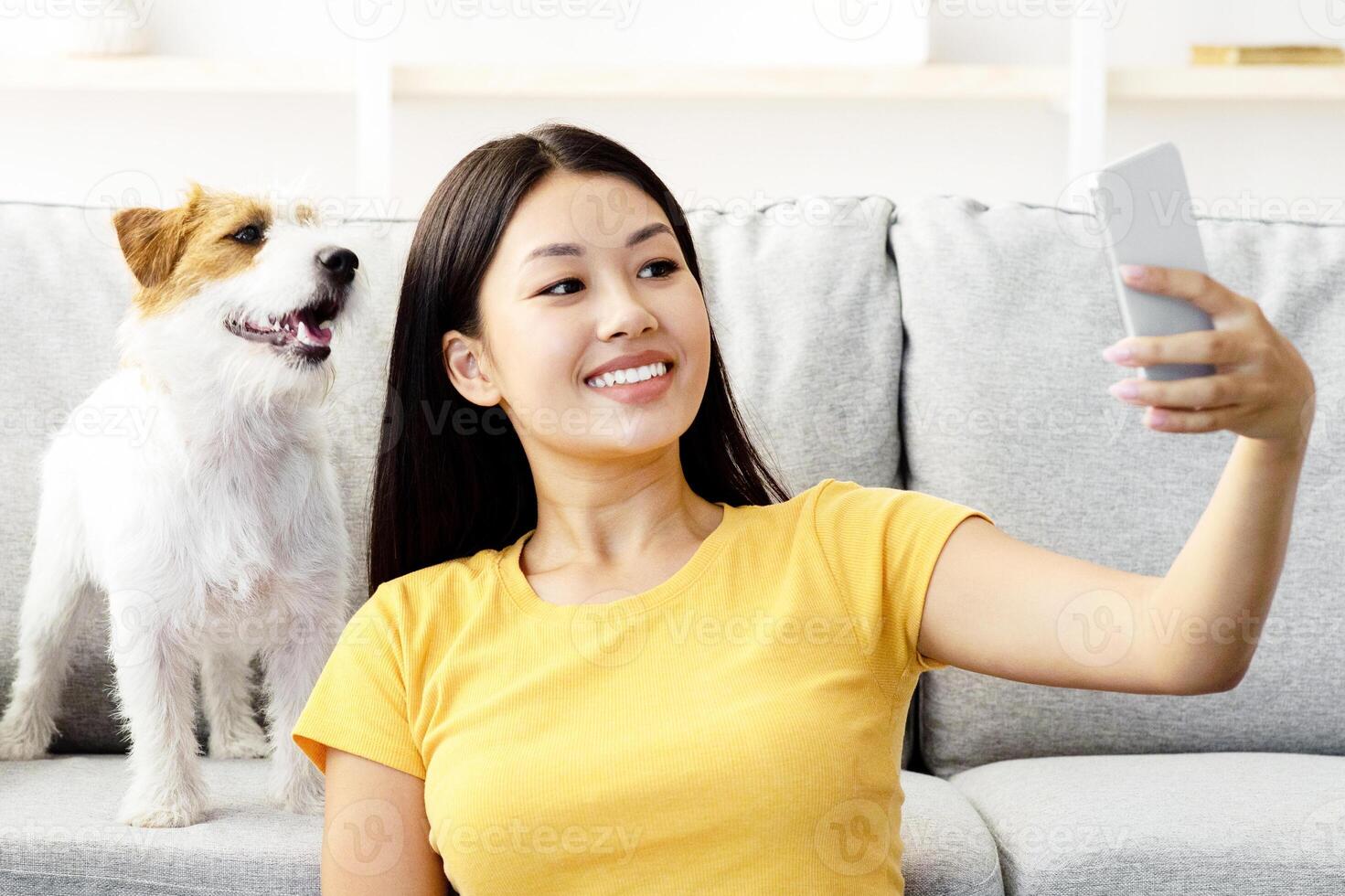 Smiling young asian woman in casual sitting by couch in living room, taking selfie with her cute fluffy dog jack russel terrier puppy while playing together at home, using cellphone, copy space photo