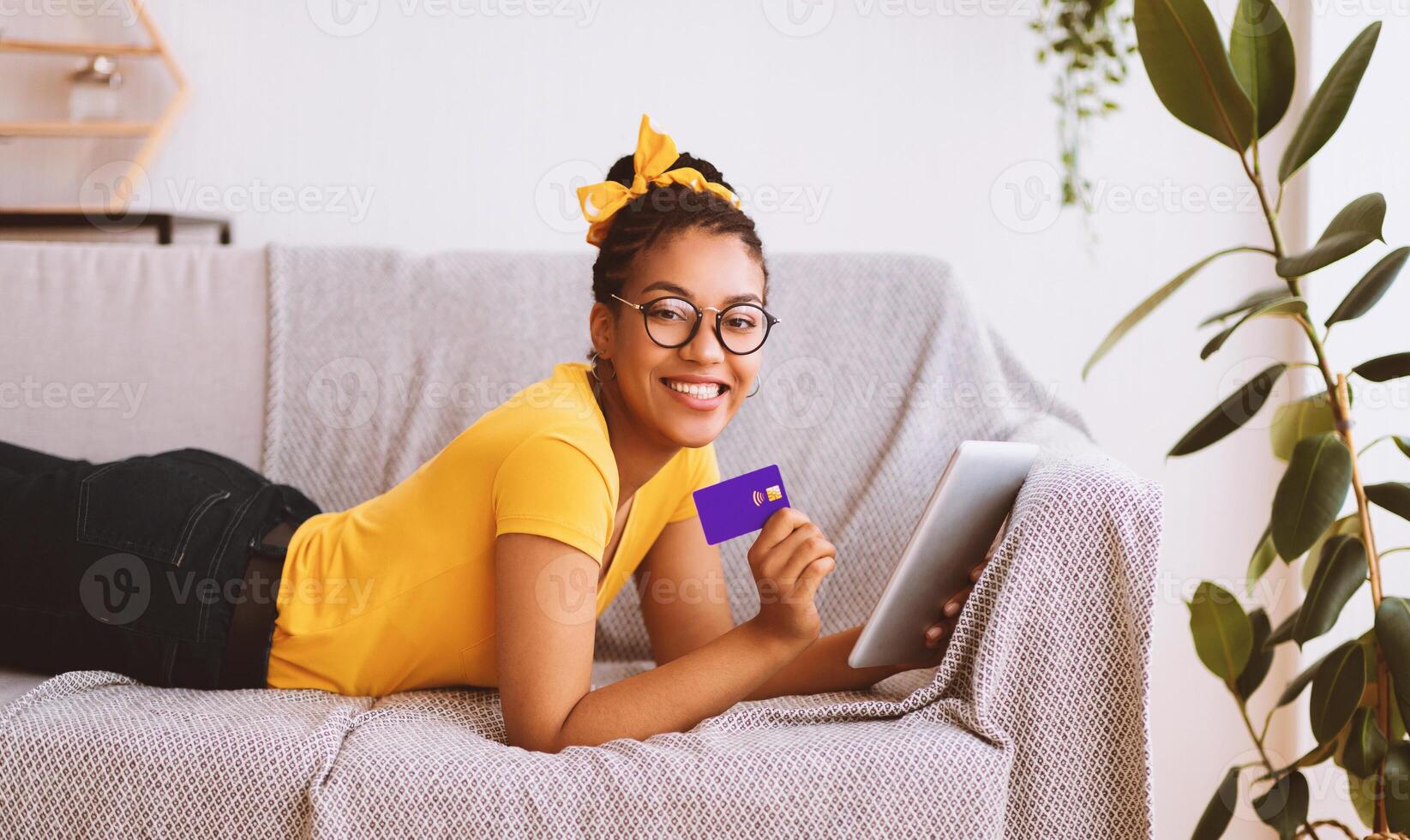 Easy Online Payment Concept. Smiling black lady in glasses using digital tablet and showing credit card, ordering purchases via internet. Woman lying on the couch and looking at camera photo