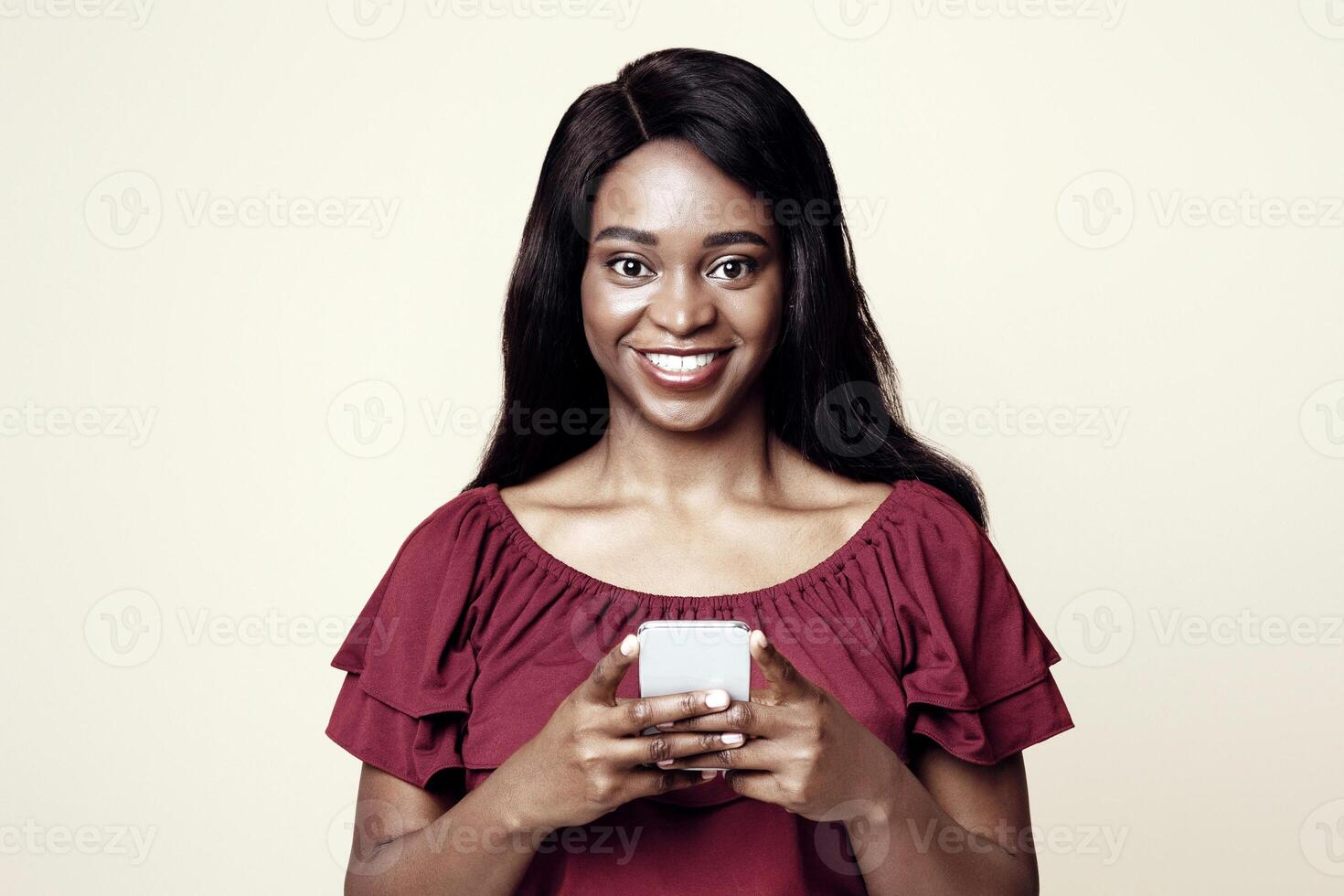 Positive young african american woman with newest cellphone looking at camera and smiling over beige studio background, using nice dating, entertaining mobile application on smartphone, copy space photo