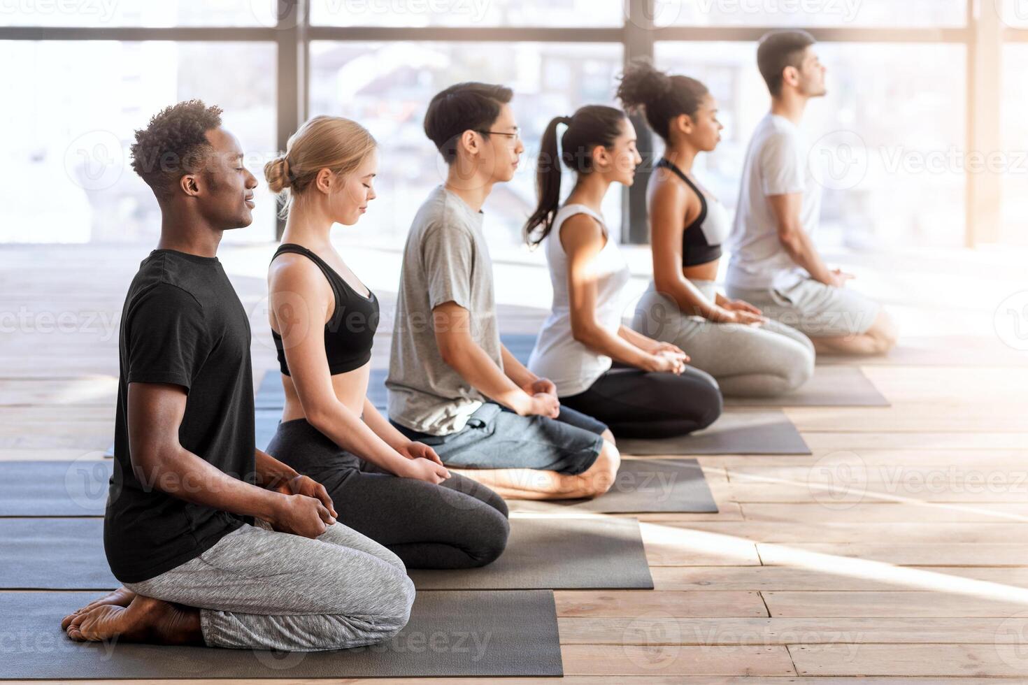 Wellness Concept. Young sporty people in yoga class making meditation exercises together, sitting on mats in modern loft studio, side view photo