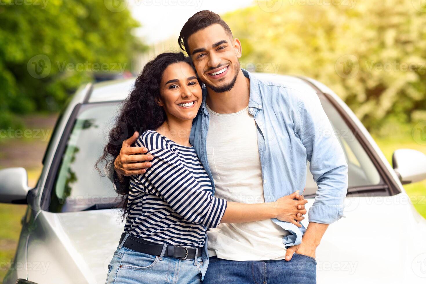 Beautiful multiethnic millennial couple having break at countryside while car trip, going vacation, standing by their nice auto, embracing and cheerfully smiling at camera, free space photo