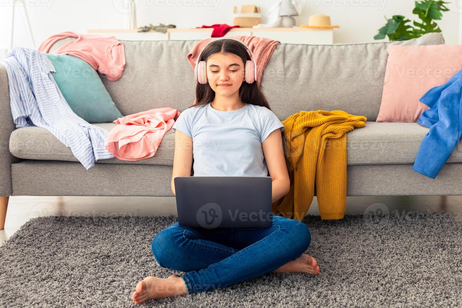 Web based schooling. Lovely Indian teen girl in headphones speaking to friends on webcam, using laptop in messy room. Cheerful adolescent using portable pc for studies or entertainment, full length photo