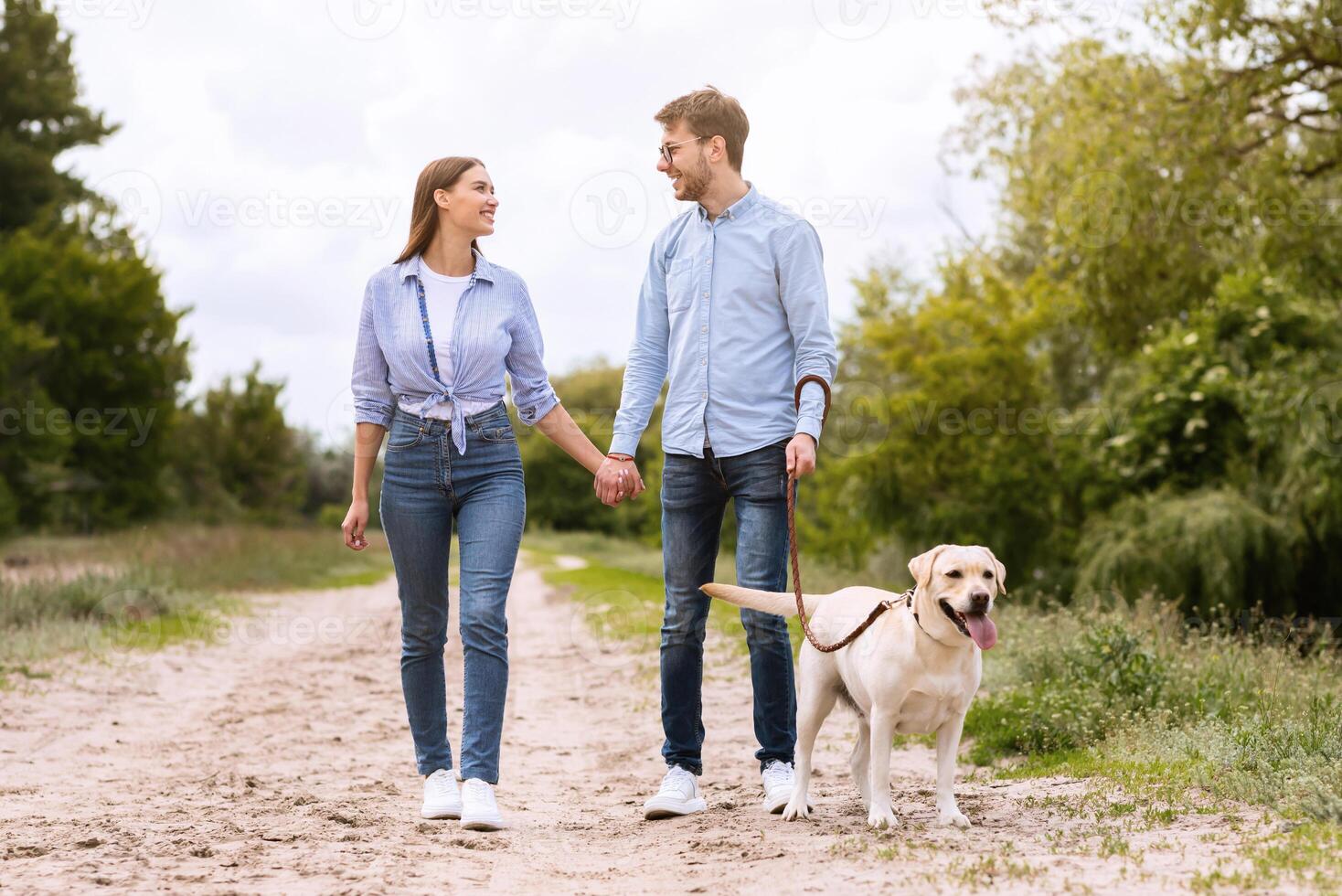Family Walk Concept. Excited young spouses walking their adorable labrador on a leash in the countryside, copy space photo