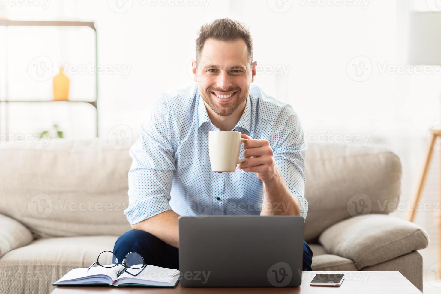 Good Morning. Smiling man drinking coffee at home and using laptop, looking at camera, copyspace photo