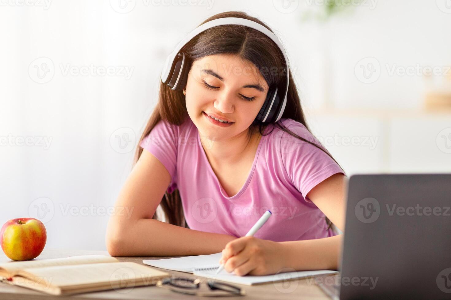 Home schooling. Joyful Indian teenage girl in headphones taking notes during online class on laptop indoors. Positive adolescent taking part in webinar, attending lecture on pc photo