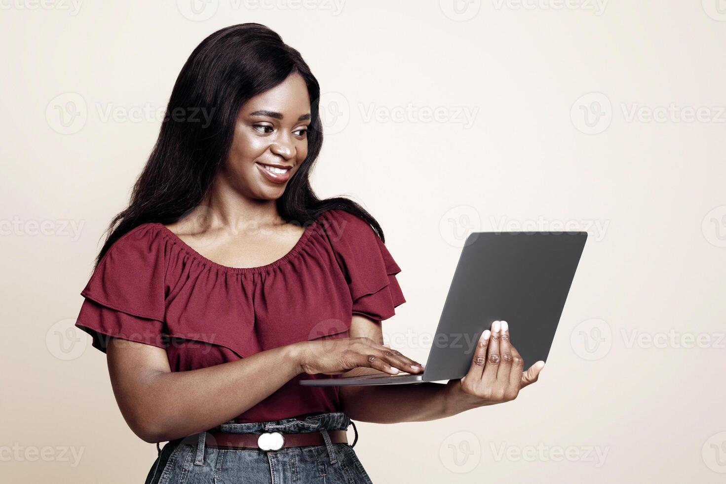 Smiling millennial black lady independent contractor working online, using modern laptop over beige studio background, typing on keyboard, studio shot, copy space. Remote job, freelance concept photo