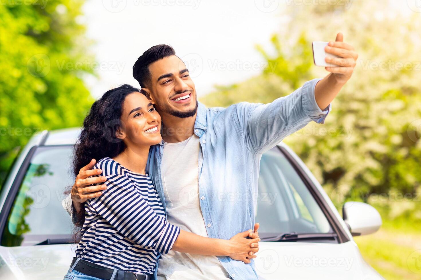 Loving mixed race couple standing by brand new car at countryside, hugging and smiling, taking selfie on mobile phone. Happy arab guy and pretty brunette lady posing by nice auto, having car trip photo