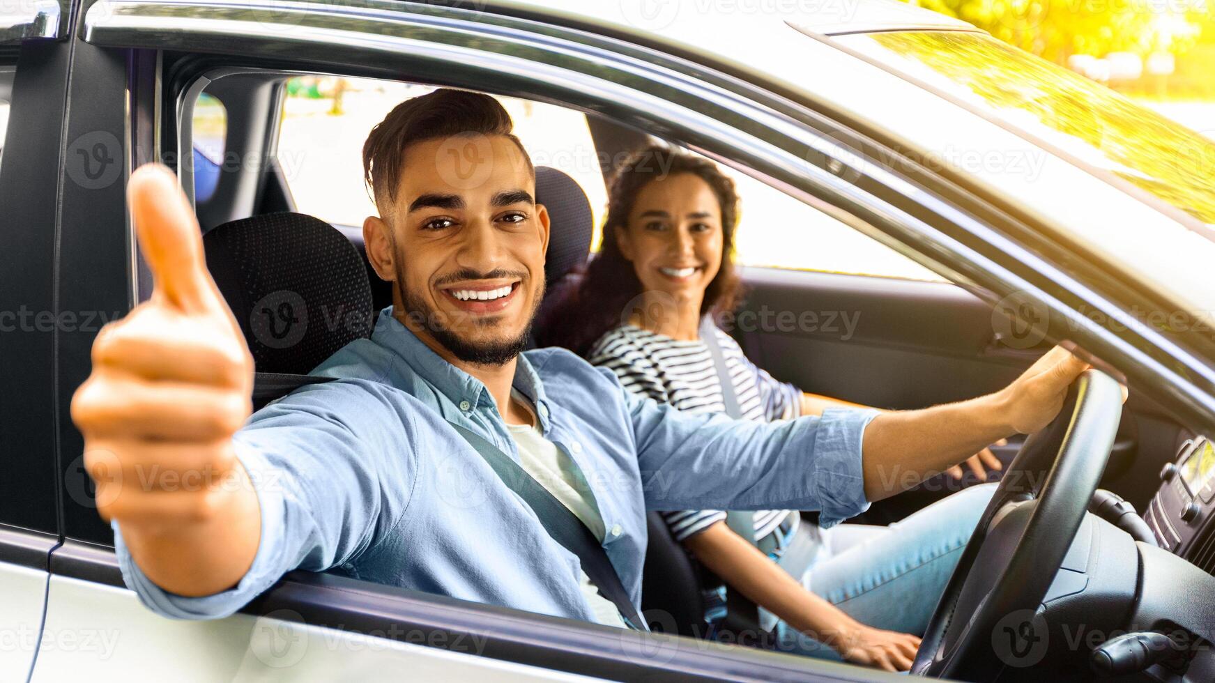 Handsome arab guy sitting inside car with gilfriend and showing thumb up, smiling at camera. Happy middle-eastern couple or family renting auto for their weekend trip, going vacation together photo