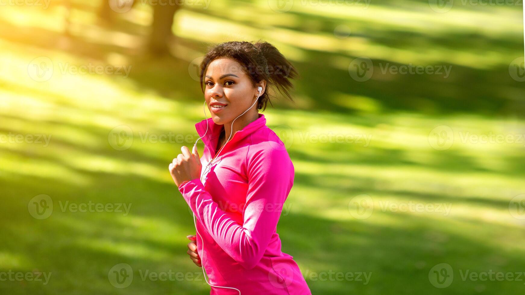 Pretty african girl in pink sweatshirt running by park, listening to music, copy space photo