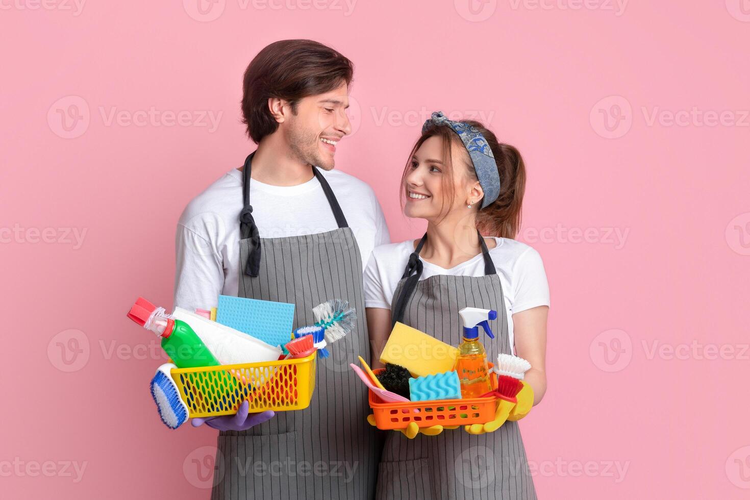 Loving young couple holding baskets with detergents and sponges, ready to make spring cleaning together, standing on pink background with free space photo