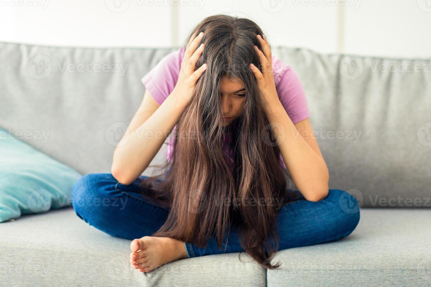 Indian teen girl sitting on sofa head in hands, absorbed in negative thoughts, having depression at home, full length. Unhappy adolescent suffering from nervous breakdown, feeling desperate photo