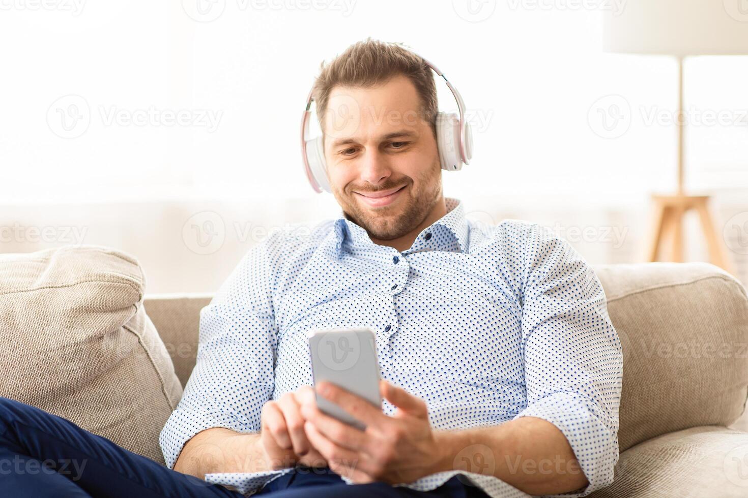 Close up portrait of serene handsome guy enjoying listening to nice song at home, sitting on sofa, holding cell phone photo