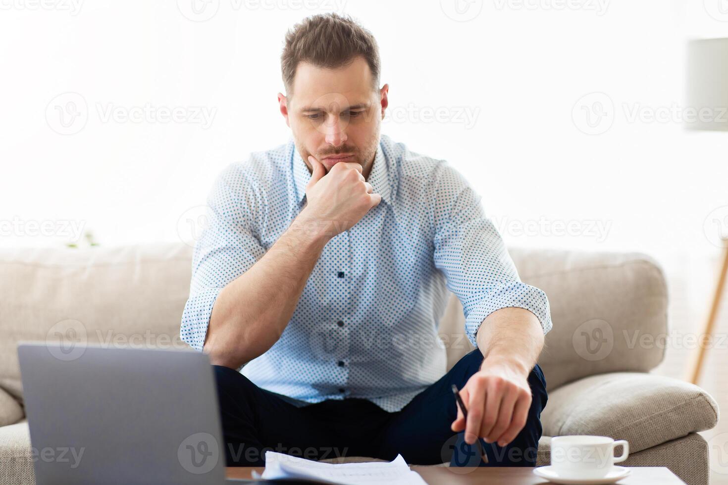Pensive young man sitting at table, looking at paper documents, working on laptop, copyspace photo
