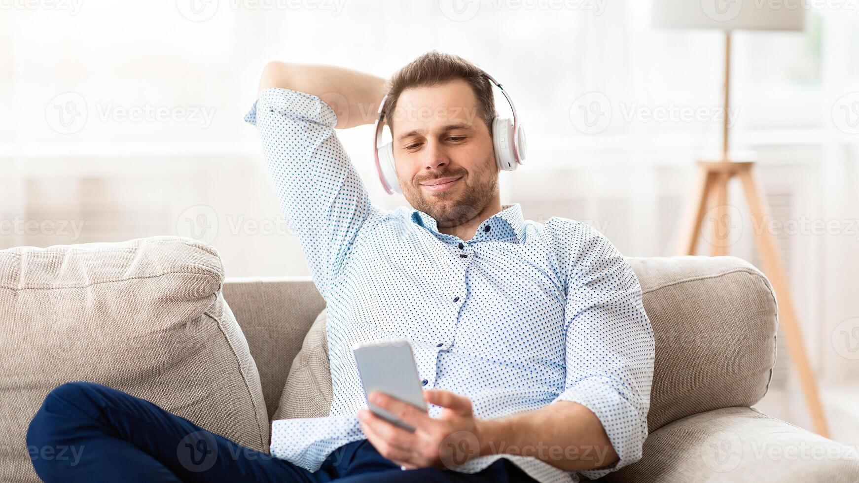 Free Time. Front view of relaxed man listening to music in wireless headphones, sitting on couch with phone, copyspace photo