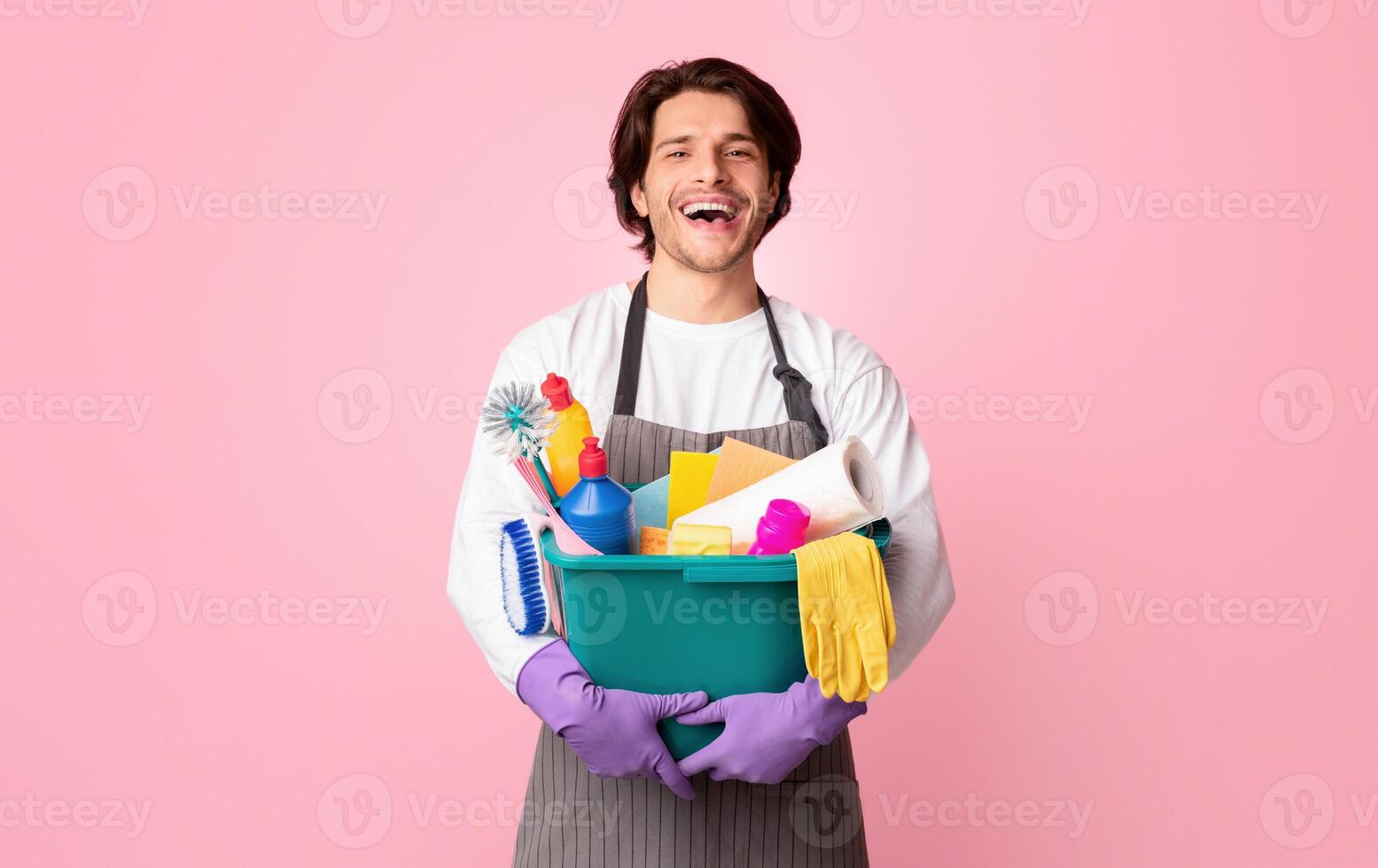 Happy young man holding bucket with cleaning supplies, posing over pink background in studio, copy space photo