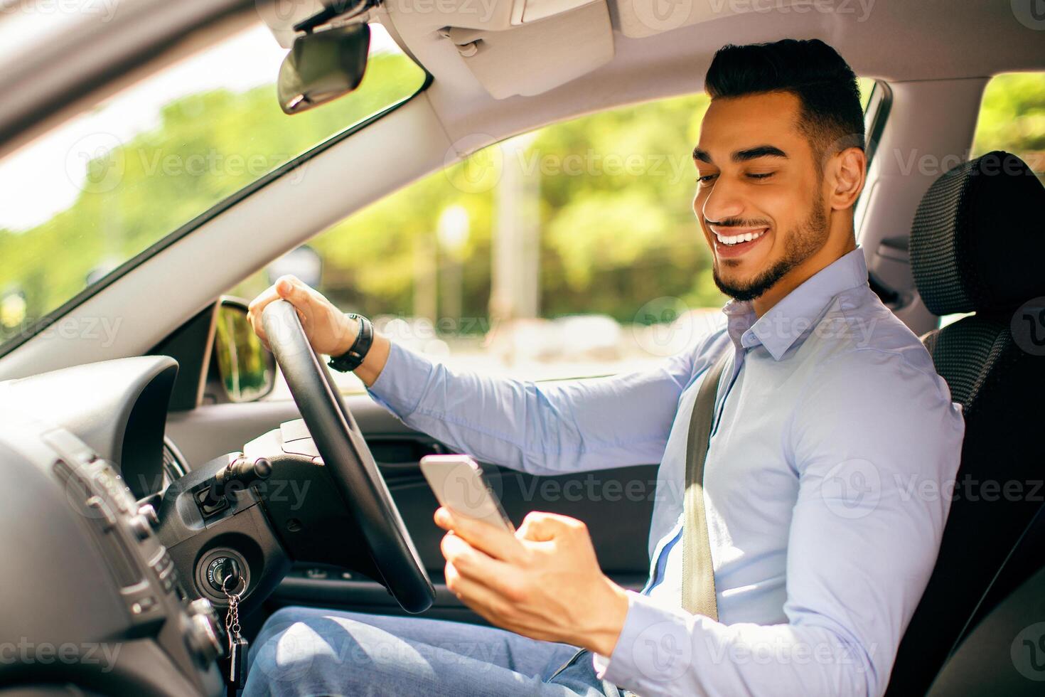 Bad driving habits, unsafe driving, driving and gadgets concept. Cheerful arab man entrepreneur going to office by auto, using cellphone while driving car, chatting with someone, side view, copy space photo