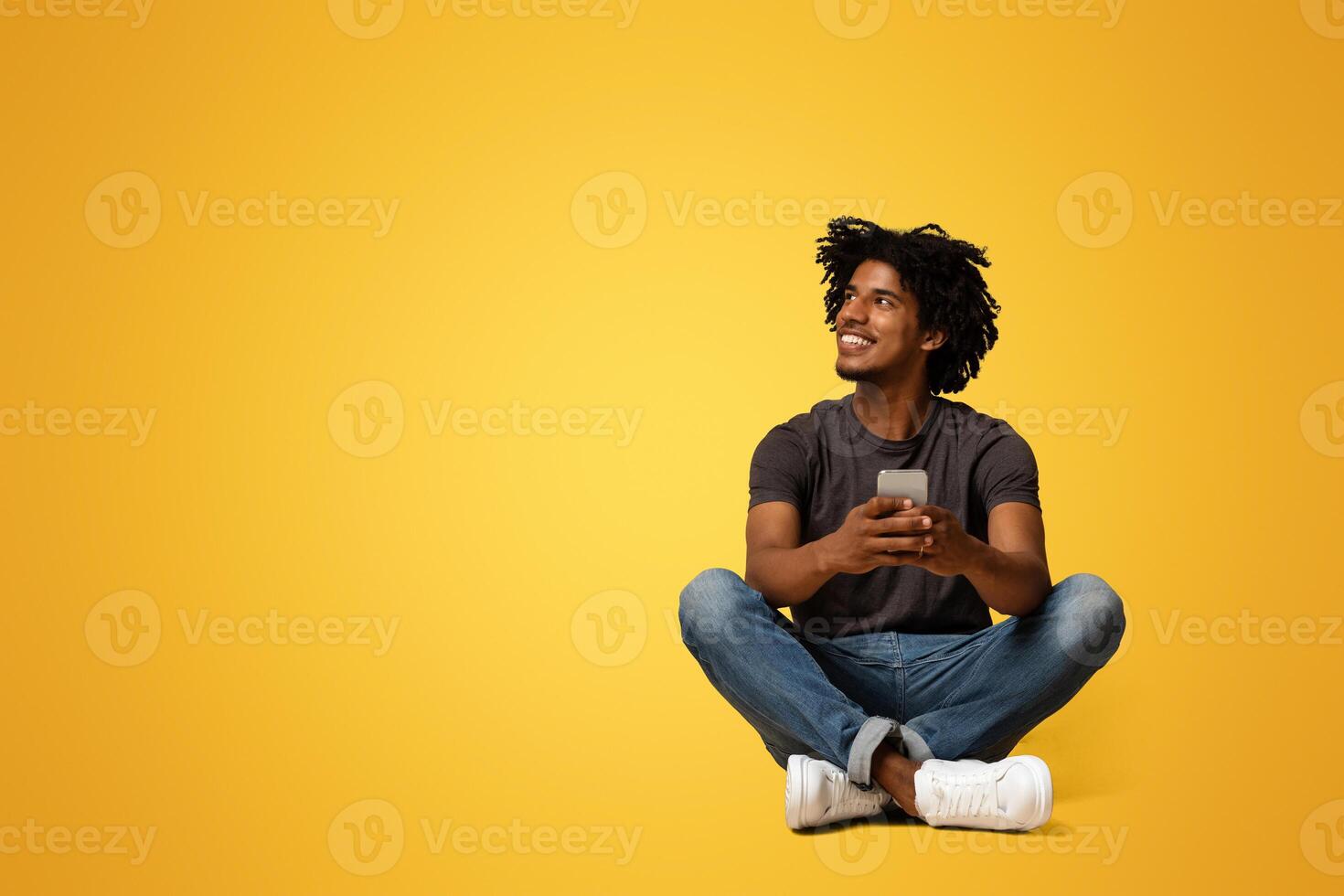 Mobile Offer. Smiling young black man with smartphone in hands sitting on floor and looking aside at copy space on beige studio background, enjoying using modern gadget for communication, panorama photo