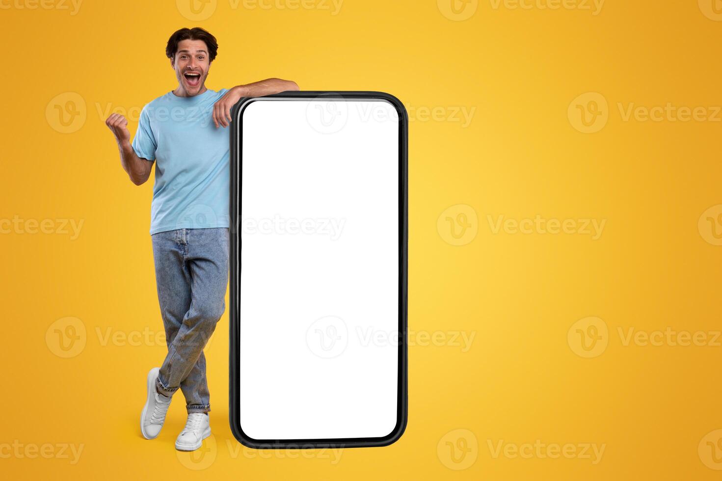 Big Luck And Win. Excited Male Leaning On Big Cell Phone With Empty White Screen Shaking Clenched Fist, Cheerful Man Celebrating Win, Standing At Orange Studio, Vertical Mock Up, Full Body Length photo
