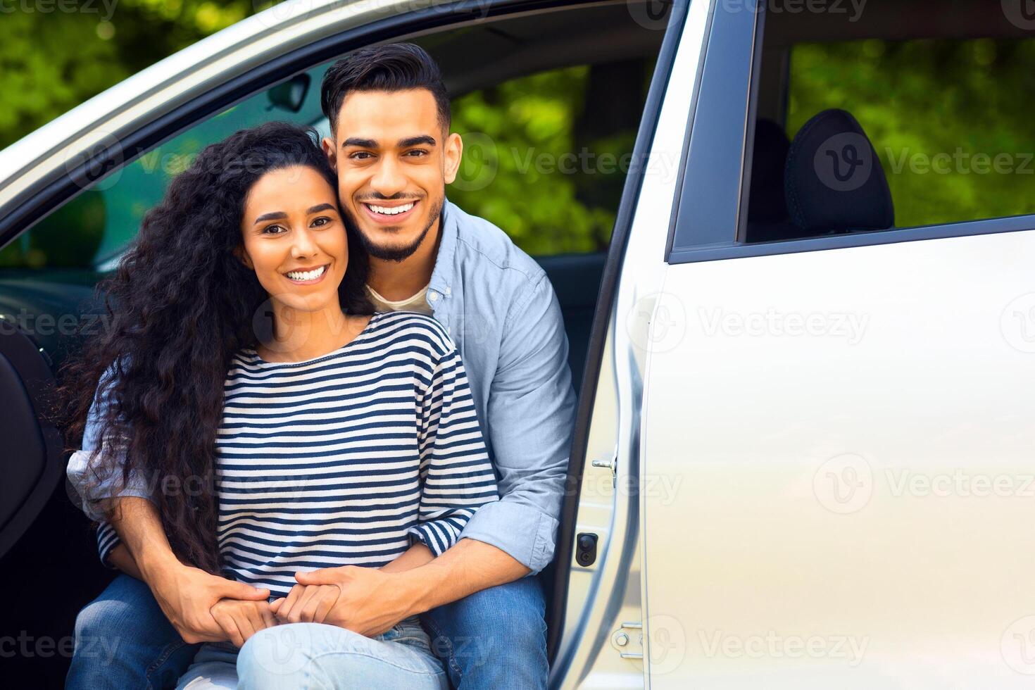 Beautiful middle-eastern lovers enjoying car trip to countryside, closeup portrait. Handsome arab guy hugging his pretty woman, sitting inside open auto, hugging and smiling at camera photo