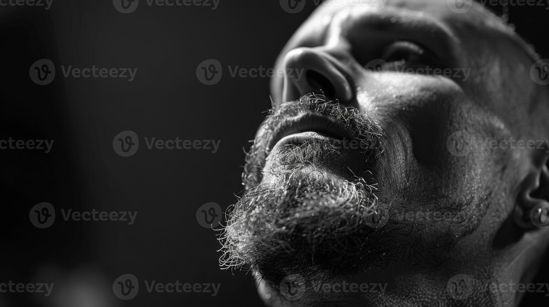 A man with a well groomed beard and intense expression looks upward, illuminated by soft light, highlighting his facial features in a moody atmosphere. photo