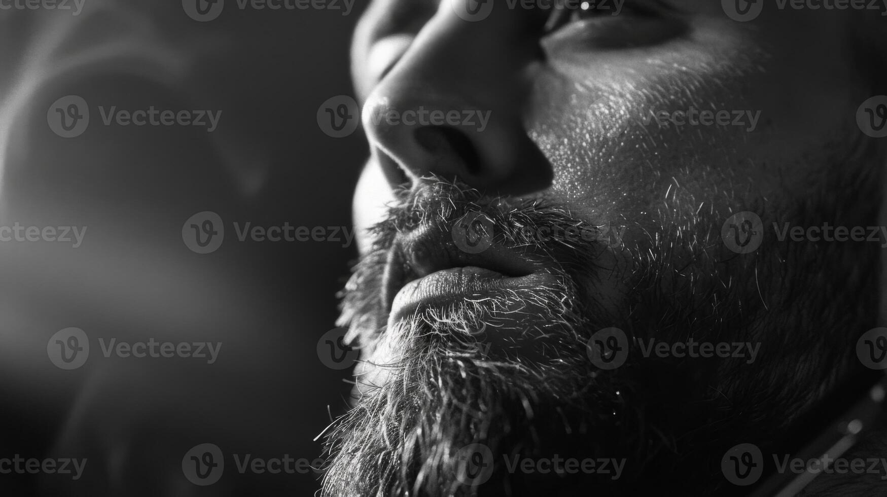 A man with a well groomed beard gazes thoughtfully with a serious expression, illuminated by soft lighting, in a relaxed and artistic environment during the evening. photo